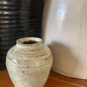 May include: A small, handcrafted ceramic vase with a rounded body and a narrow neck. The vase has a light beige colour with brown streaks and spots, giving it a rustic appearance. The vase is sitting on a wooden surface.
