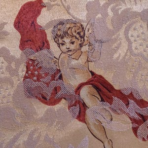 K&ouml;nnte beinhalten: Nahaufnahme eines gewebten Stoffes mit einem Cherub-Design. Der Cherub ist mit heller Haut, lockigem Haar und einem roten Tuch dargestellt. Der Hintergrund hat ein Blumenmuster in Beige und Hellviolett.