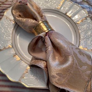 K&ouml;nnte beinhalten: Eine gefaltete, gold-beige gemusterte Serviette wird von einem geh&auml;mmerten goldenen Serviettenring gehalten und liegt auf einem wei&szlig;en Teller mit goldenem Rand. Der Teller steht auf einem transparenten Untersetzer und einer rot-wei&szlig; gestreiften Tischdecke.