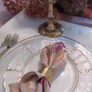 K&ouml;nnte beinhalten: Elegante Tischdekoration mit einem wei&szlig;en Teller mit Goldrand und einer gemusterten Serviette, die von einem goldenen Ring gehalten wird. Ein goldener Kerzenst&auml;nder und Trockenblumen schaffen ein edles Ambiente.