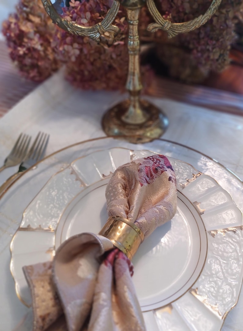 K&ouml;nnte beinhalten: Elegante Tischdekoration mit einem wei&szlig;en Teller mit Goldrand und einer gemusterten Serviette, die von einem goldenen Ring gehalten wird. Ein goldener Kerzenst&auml;nder und Trockenblumen schaffen ein edles Ambiente.