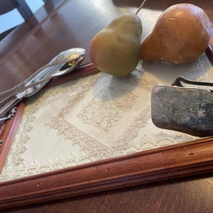 Peut inclure: Nature morte avec un plateau encadré en bois et un motif de dentelle crème. Deux poires et deux ustensiles de service en argent sont placés sur le plateau. Les ustensiles ont des poignées ornées.