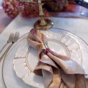 K&ouml;nnte beinhalten: Elegante Tischdekoration mit einem wei&szlig;en Teller mit Goldrand, einer gefalteten Blumenserviette, die von einem goldenen Ring gehalten wird, und einem goldenen Kerzenleuchter. Das Arrangement umfasst einen transparenten Glasuntersetzer und eine silberne Gabel.