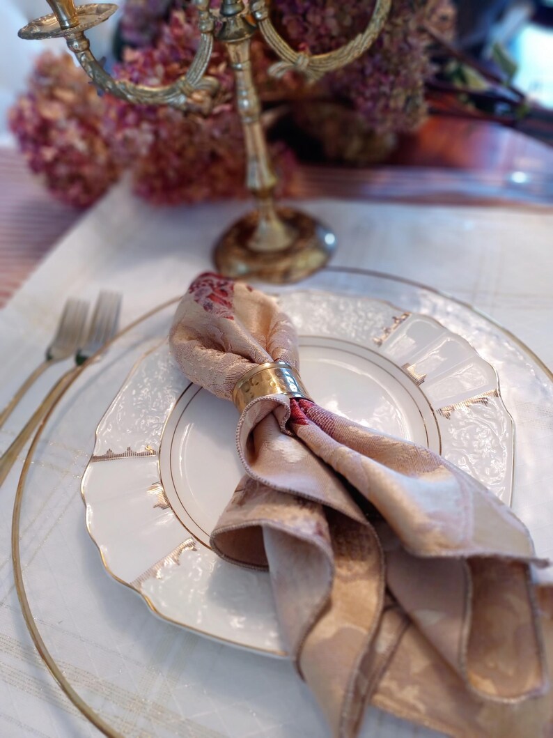 K&ouml;nnte beinhalten: Elegante Tischdekoration mit einem wei&szlig;en Teller mit Goldrand, einer gefalteten Blumenserviette, die von einem goldenen Ring gehalten wird, und einem goldenen Kerzenleuchter. Das Arrangement umfasst einen transparenten Glasuntersetzer und eine silberne Gabel.