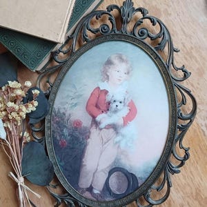 Puede incluir: Un marco de fotos ovalado antiguo con un retrato vintage de un niño sosteniendo un perro blanco pequeño. El marco es ornamentado, con un acabado en bronce oscuro. El retrato presenta a un niño con una chaqueta roja y pantalones claros. Flores secas y libros en el fondo.