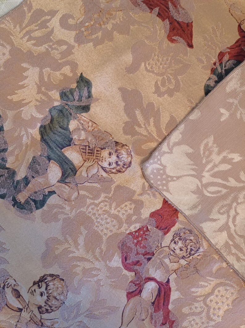 K&ouml;nnte beinhalten: Nahaufnahme eines beigefarbenen Stoffes mit einem sich wiederholenden Muster aus Cherubinen und floralen Designs. Die Cherubinen sind in verschiedenen Posen dargestellt, mit Details in Gr&uuml;n, Rot und Beige. Der Stoff hat ein strukturiertes Aussehen.