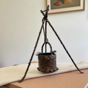 May include: A rustic, bronze-colored miniature well sculpture. The well is supported by three twig-like legs, with a small bucket suspended from above. The sculpture is placed on a stack of books and a cream-colored cloth.