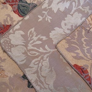 K&ouml;nnte beinhalten: Nahaufnahme eines Abschnitts eines floralen Wandteppichs. Der Stoff weist ein sich wiederholendes Muster aus gro&szlig;en, stilisierten Blumen in Creme und Beige mit Akzenten in Rot, Gr&uuml;n und Braun auf. Die Kanten sind mit einem kontrastierenden Rand versehen.