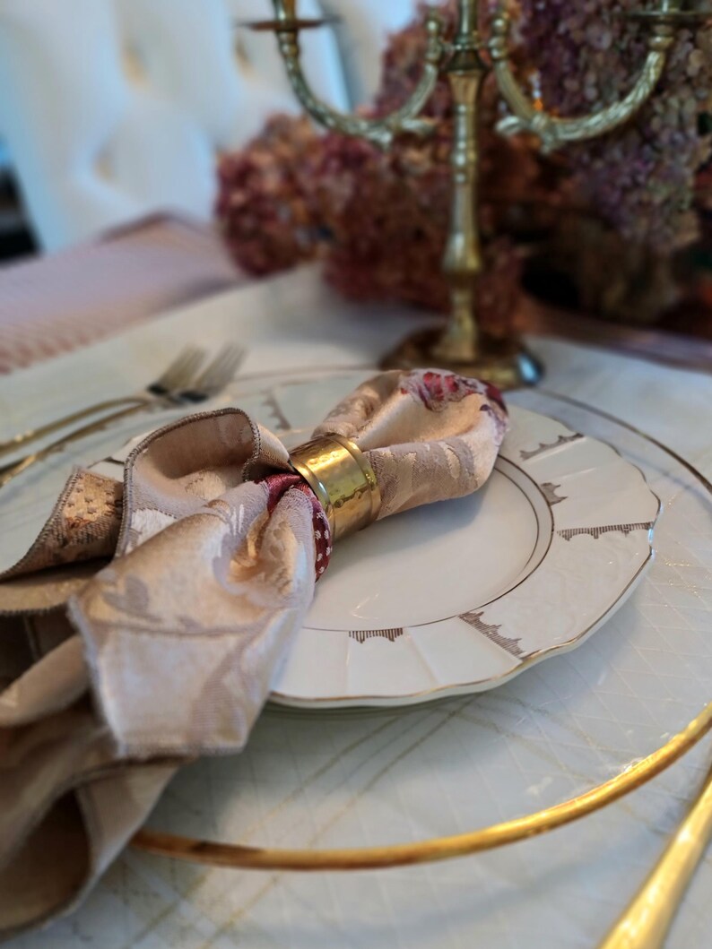 K&ouml;nnte beinhalten: Tischarrangement mit einem goldenen Serviettenring, der eine beige und floral gemusterte Serviette h&auml;lt. Ein wei&szlig;er Teller mit Goldrand liegt auf einem transparenten Untersetzer. Ein goldener Kerzenst&auml;nder und Besteck befinden sich im Hintergrund.