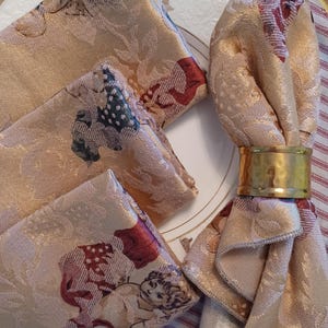 K&ouml;nnte beinhalten: Set aus vier beigefarbenen Damastservietten mit einem Cherub- und Blumenmuster, kombiniert mit einem goldenen Serviettenring. Die Servietten sind auf einem wei&szlig;en Teller mit Goldrand drapiert.
