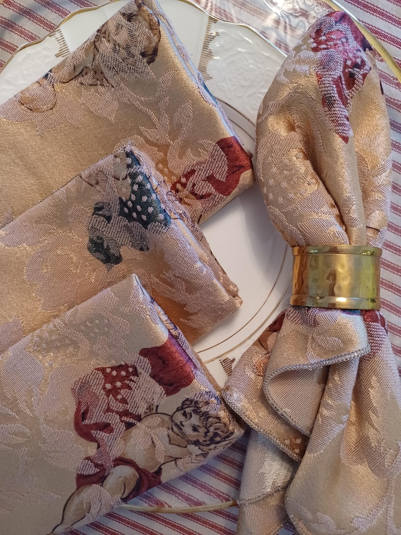 K&ouml;nnte beinhalten: Set aus vier beigefarbenen Damastservietten mit einem Cherub- und Blumenmuster, kombiniert mit einem goldenen Serviettenring. Die Servietten sind auf einem wei&szlig;en Teller mit Goldrand drapiert.