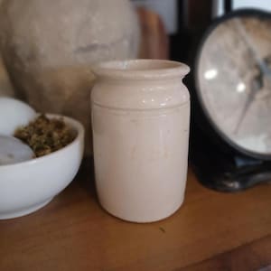May include: A small, off-white ceramic jar with a cylindrical body and a slightly flared rim. The jar has a simple, classic design and appears to be vintage. Other items in the background include a mortar and pestle, and a scale.