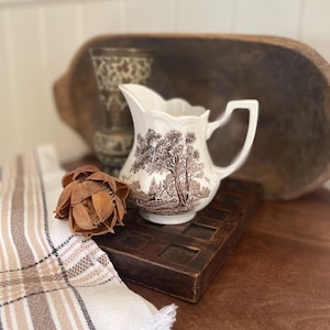 Peut inclure: Une cruche en céramique blanche avec un motif de scène pastorale marron. La cruche est posée sur un carré en bois foncé, à côté d'une fleur séchée et d'un tissu à rayures et à carreaux. Un vase décoratif et un bol en bois sont en arrière-plan.