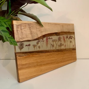May include: Wooden serving board with a natural edge and a clear resin inlay. The resin section features dried flowers in shades of pink, brown, and beige. The board is rectangular with a diagonal cut on one corner.