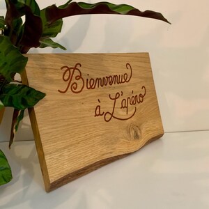 May include: A wooden sign with the words "Bienvenue à L'apéro" in red script. The sign has a natural wood grain and a live edge. A green plant is visible in the background.