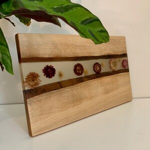 May include: Wooden cutting board with a clear resin inlay. The inlay features dried flowers in shades of red, yellow, and pink. The board is made of light-colored wood with a natural grain pattern. The board is rectangular and has a smooth surface.