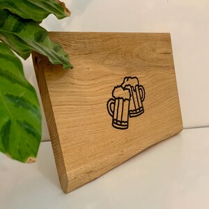 May include: A light-colored wooden cutting board with a black outline of two beer mugs. The board has a natural wood grain and a slightly angled edge. The design is simple and rustic.