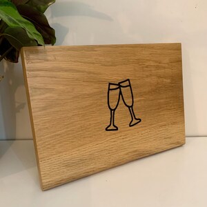 May include: A rectangular wooden sign with a light brown finish. The sign features a black outline of two champagne glasses clinking together. The sign is displayed upright, suggesting it is meant to stand on a surface.