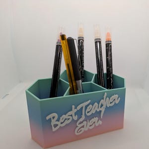 May include: A hexagonal desk organiser with a gradient design in pink, light blue, and dark blue. The organiser has six compartments and says "Best Teacher Ever" in white lettering. The organiser is filled with pens and markers.