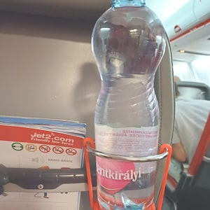 May include: A clear plastic water bottle with a pink cap and blue neck is held in an orange holder. The bottle has a label with text. The holder is attached to an airplane seat. A Jet2.com brochure is visible.