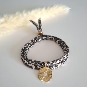 Könnte beinhalten: Ein Armband mit Leopardenmuster und einem goldfarbenen Anhänger. Das Armband besteht aus einem weichen Stoffmaterial mit einem braunen und schwarzen Tiermuster. Der Anhänger ist ein goldfarbener, sonnenförmiger Charm.