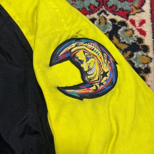 May include: A close-up of a yellow and black jacket. The jacket features a colorful crescent moon patch with a smiling face, stars, and vibrant accents. The jacket's design includes a black panel on the left side.
