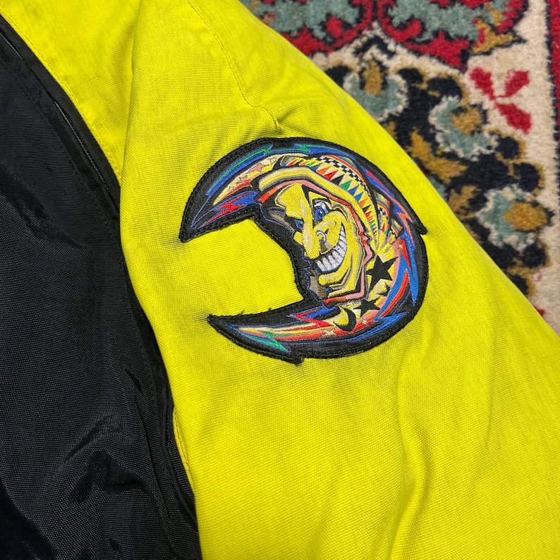 May include: A close-up of a yellow and black jacket. The jacket features a colorful crescent moon patch with a smiling face, stars, and vibrant accents. The jacket's design includes a black panel on the left side.