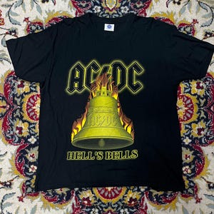 Könnte beinhalten: Schwarzes T-Shirt mit dem AC/DC-Logo in Gelb, einer brennenden Glocke und dem Text "HELL'S BELLS". Das T-Shirt besteht aus einem weichen Material. Ein klassisches Rockband-Merchandise.