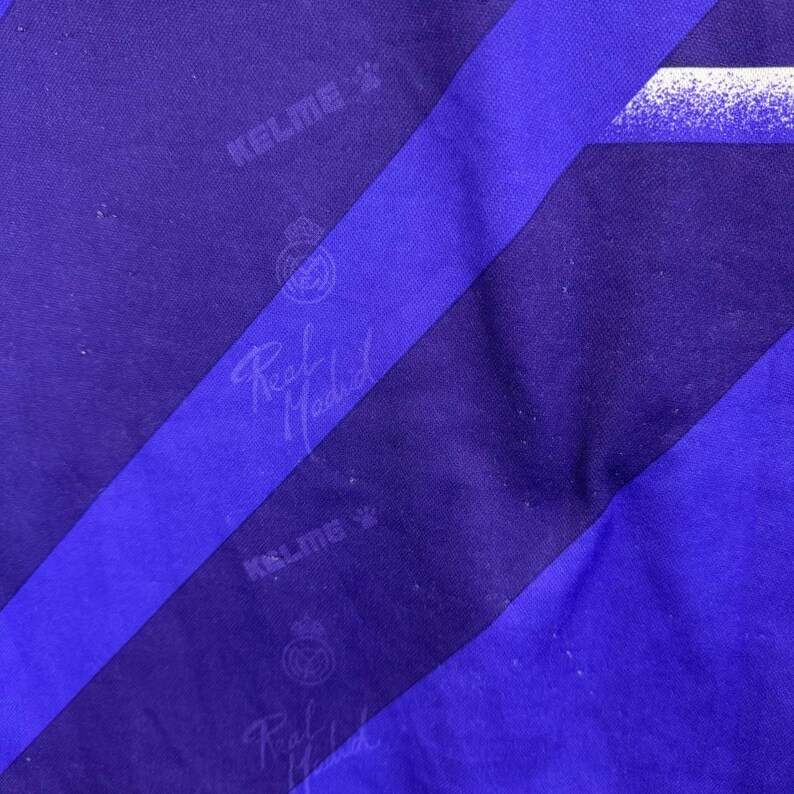 Puede incluir: Primer plano de una tela morada y azul con rayas diagonales. La tela presenta el texto "KELME" y "Real Madrid" en blanco. Tambi&eacute;n es visible el logotipo del Real Madrid.