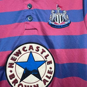 Peut inclure: Gros plan sur un maillot de rugby ray&eacute; rose et bleu. Le maillot pr&eacute;sente l'&eacute;cusson de Newcastle United et un &eacute;cusson "Brown Ale". Le maillot a un col boutonn&eacute; et un style rugby classique.