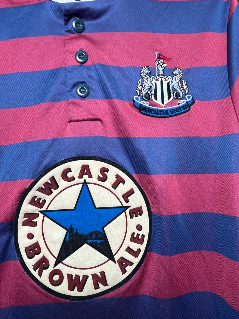 Peut inclure: Gros plan sur un maillot de rugby ray&eacute; rose et bleu. Le maillot pr&eacute;sente l'&eacute;cusson de Newcastle United et un &eacute;cusson "Brown Ale". Le maillot a un col boutonn&eacute; et un style rugby classique.