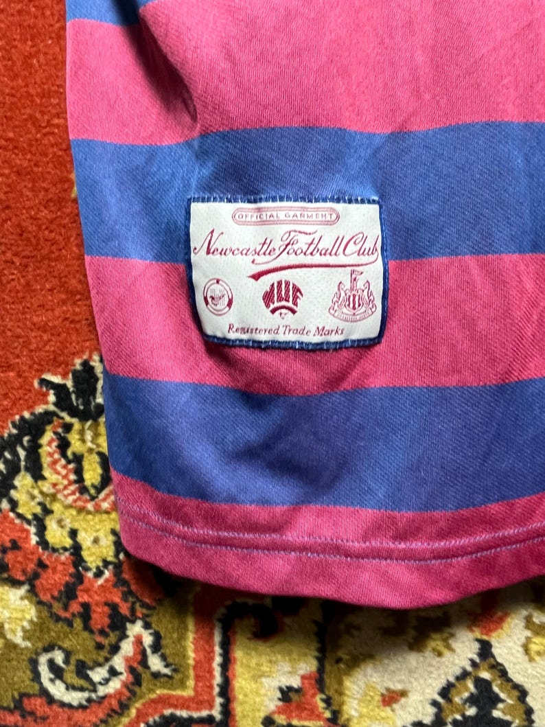 Peut inclure: Gros plan sur un t-shirt &agrave; rayures roses et bleues. Le t-shirt porte une &eacute;tiquette avec l'inscription "Newcastle Football Club", ainsi que les mentions "Official Garment" et "Registered Trade Marks".