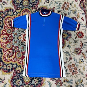 Puede incluir: Una camiseta azul de manga corta con cremallera en el cuello. La camiseta tiene rayas blancas, rojas y negras en los laterales y en las mangas. El cuello está adornado con rayas blancas, rojas y negras.