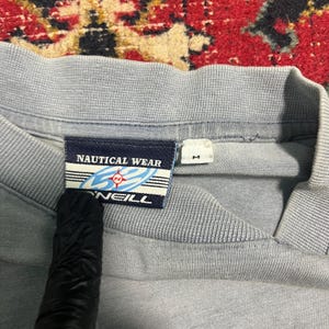 May include: Close-up of a pale blue shirt collar with a tag that reads "NAUTICAL WEAR" and "O'NEILL". The tag features a nautical design with a compass rose. A size "M" tag is also visible.