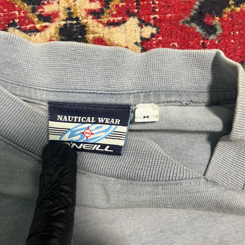 May include: Close-up of a pale blue shirt collar with a tag that reads "NAUTICAL WEAR" and "O'NEILL". The tag features a nautical design with a compass rose. A size "M" tag is also visible.