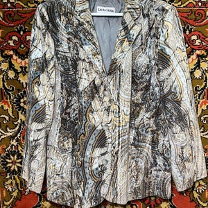 May include: A patterned blazer with a grey lining and three buttons. The blazer features a swirling design in shades of beige, brown, and blue. The label reads "LAURA LEBEK". The blazer is hanging on a white hanger.