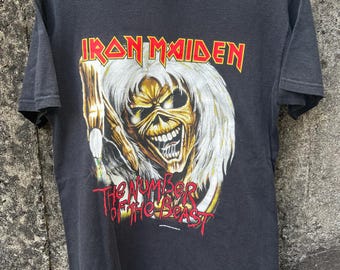 May include: Black t-shirt featuring the band name "Iron Maiden" in yellow and red lettering above a graphic of a skeletal figure with white hair. The text "The Number of the Beast" is below the graphic in red.