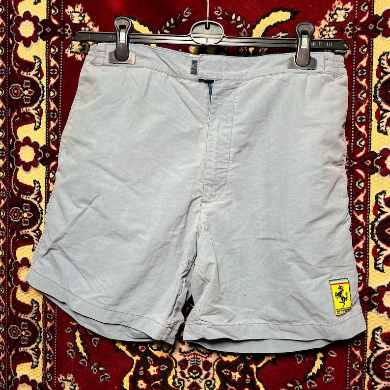Puede incluir: Pantalones cortos azul claro con cierre de cremallera y un peque&ntilde;o parche con el logotipo de Ferrari en la parte inferior de la pierna derecha. Los pantalones cortos son de material ligero y cuelgan de una percha negra.