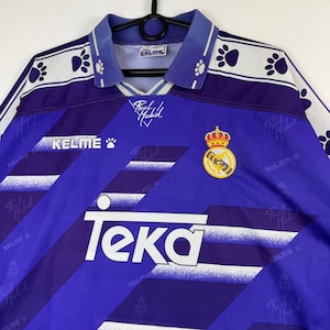 Puede incluir: Una camiseta de f&uacute;tbol vintage morada y blanca con el texto "Teka" y "Kelme". La camiseta presenta un escudo con una corona y las palabras "Real Madrid". El cuello y las mangas tienen un estampado de huellas.