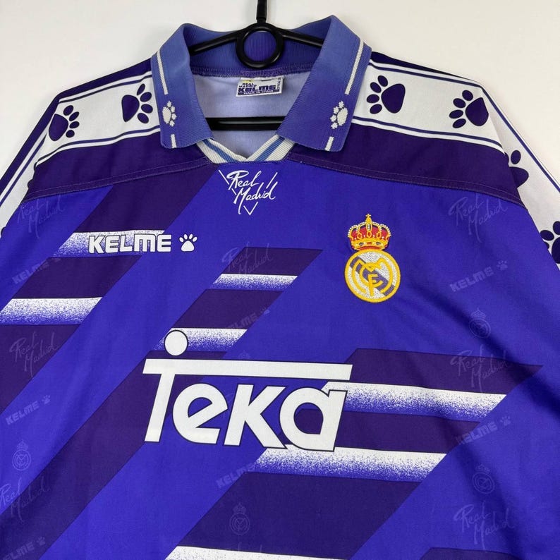 Puede incluir: Una camiseta de f&uacute;tbol vintage morada y blanca con el texto "Teka" y "Kelme". La camiseta presenta un escudo con una corona y las palabras "Real Madrid". El cuello y las mangas tienen un estampado de huellas.