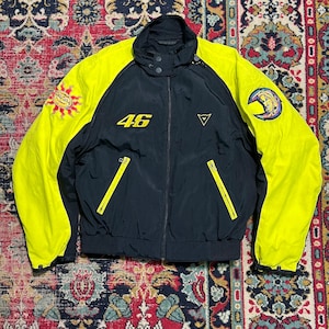 May include: Black and yellow motorcycle jacket with the number 46 in yellow on the chest. The jacket has a stand-up collar, two zippered pockets, and patches on the sleeves. The sleeves and shoulders are yellow, and the body is black.