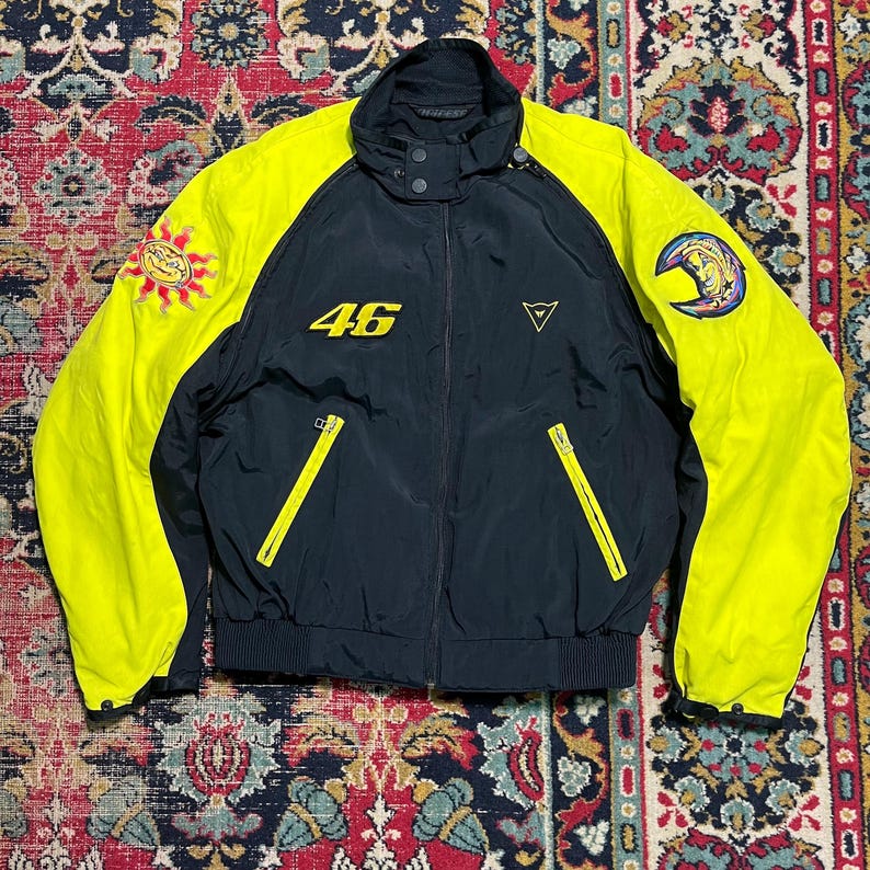 May include: Black and yellow motorcycle jacket with the number 46 in yellow on the chest. The jacket has a stand-up collar, two zippered pockets, and patches on the sleeves. The sleeves and shoulders are yellow, and the body is black.