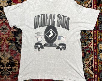 May include: A light gray t-shirt featuring the text "WHITE SOX" in a stylized font. Below the text is a baseball diamond logo with a sock, and the word "CHICAGO" on a banner. Additional logos are also present.