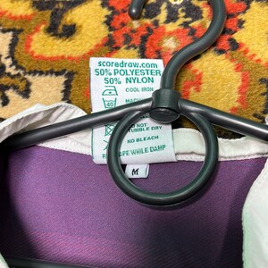 Peut inclure: Une chemise &agrave; col violette et blanche avec une &eacute;tiquette indiquant "scoredraw.com 50% POLYESTER 50% NYLON". La chemise est suspendue &agrave; un cintre noir. L'&eacute;tiquette comprend &eacute;galement des instructions de lavage.