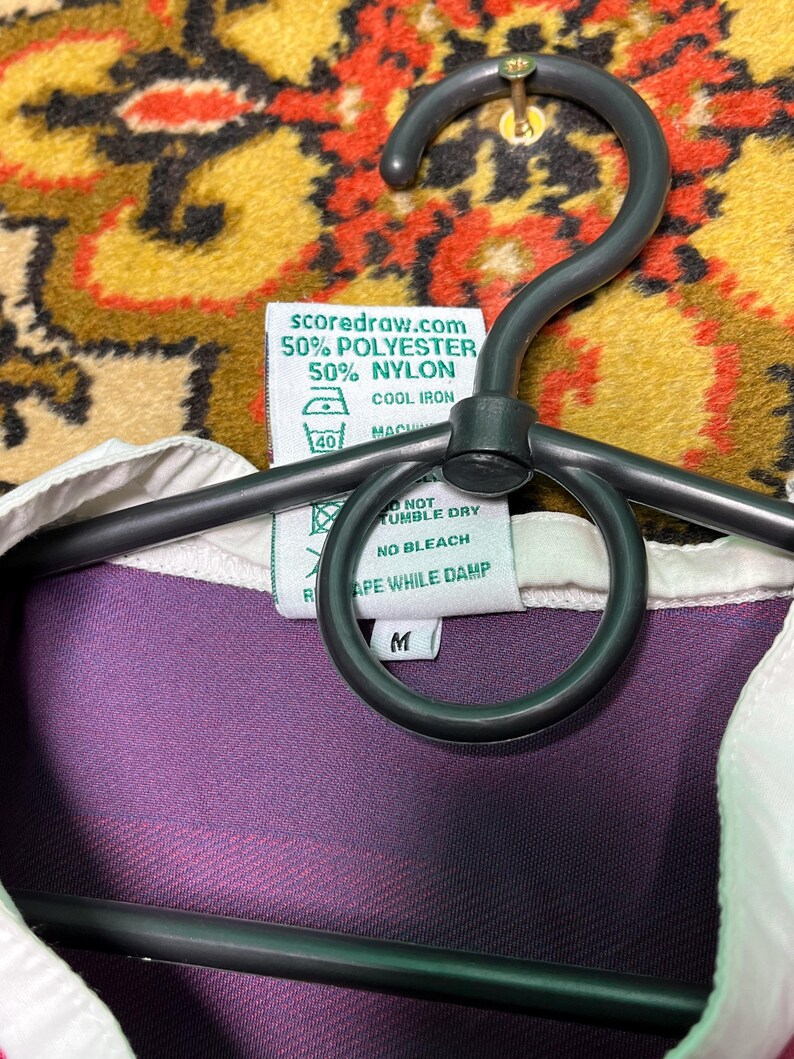 Peut inclure: Une chemise &agrave; col violette et blanche avec une &eacute;tiquette indiquant "scoredraw.com 50% POLYESTER 50% NYLON". La chemise est suspendue &agrave; un cintre noir. L'&eacute;tiquette comprend &eacute;galement des instructions de lavage.