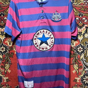 Peut inclure: Un maillot ray&eacute; rose et bleu avec l'&eacute;cusson de Newcastle United et le logo "Newcastle Brown Ale". Le maillot a un col boutonn&eacute; et une petite &eacute;tiquette. Manches courtes.