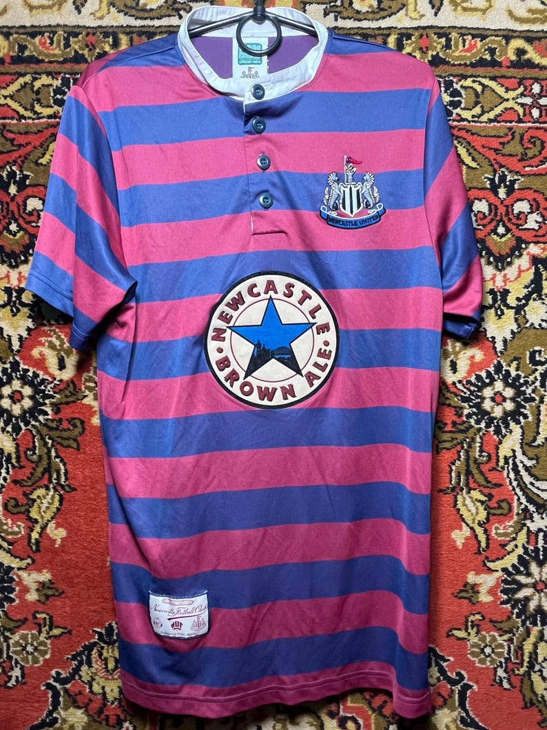 Peut inclure: Un maillot ray&eacute; rose et bleu avec l'&eacute;cusson de Newcastle United et le logo "Newcastle Brown Ale". Le maillot a un col boutonn&eacute; et une petite &eacute;tiquette. Manches courtes.