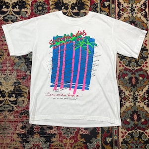 Può includere: T-shirt bianco con un design grafico colorato. Il design presenta il testo "COCOMANGAS SHOOTER BAR" in rosso e verde, insieme a un elenco di bevande e la frase "STILL STANDING AFTER 15... DO IT FOR YOUR COUNTRY."