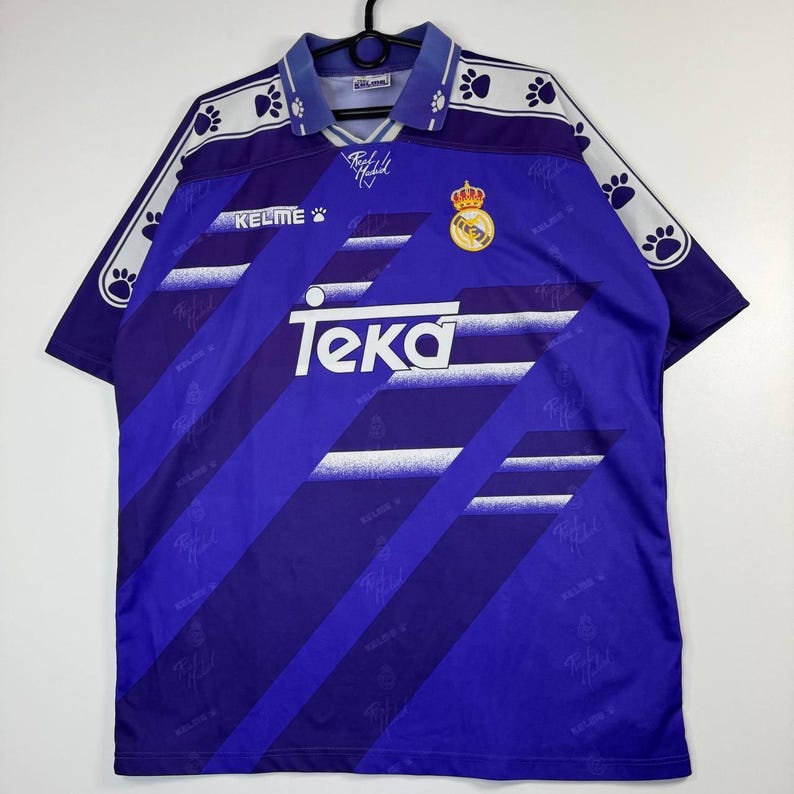 Puede incluir: Una camiseta de f&uacute;tbol morada vintage con detalles blancos y azules. La camiseta presenta el logo "Teka", un escudo del equipo y el logo "Kelme". Las mangas tienen un dise&ntilde;o de huellas de patas. La camiseta est&aacute; colgada en una percha negra.