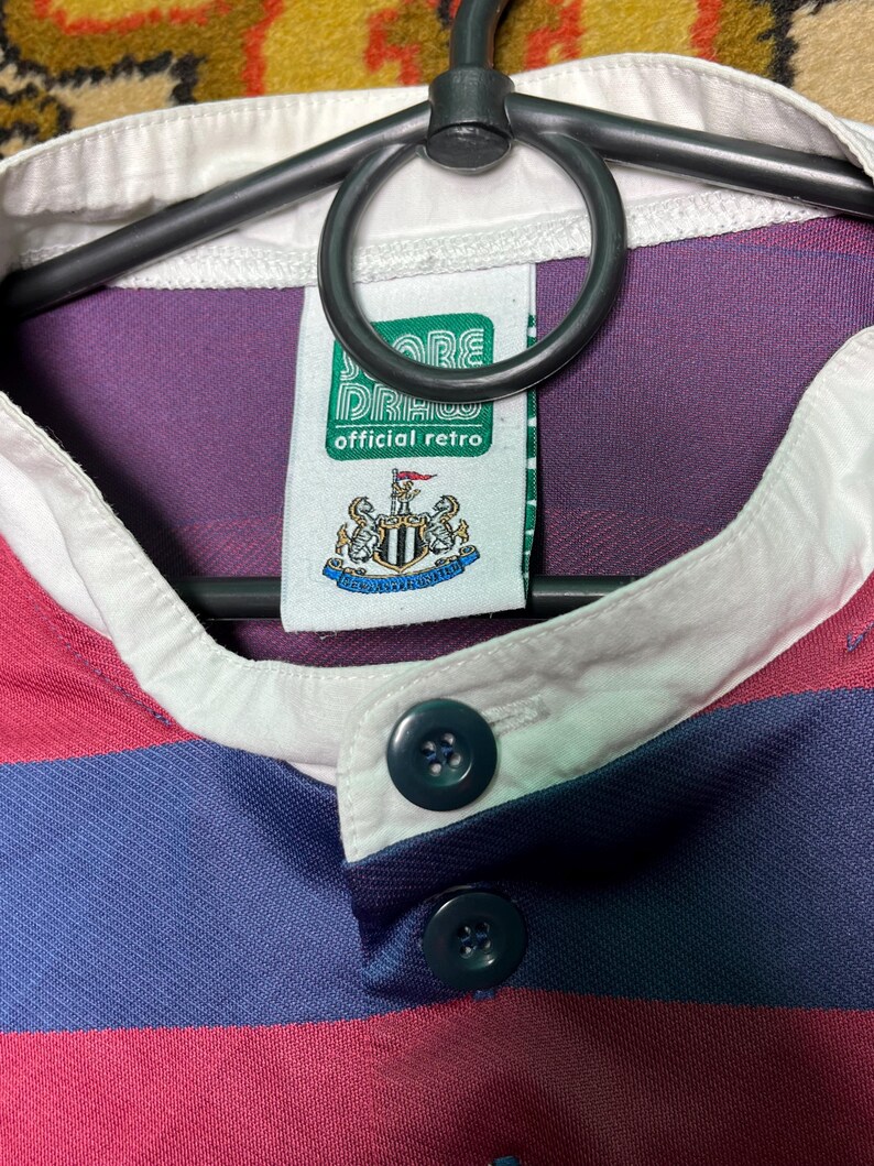 Peut inclure: Gros plan sur un maillot de rugby r&eacute;tro avec un col blanc et une patte de boutonnage. Le maillot pr&eacute;sente des rayures horizontales rouges et bleu marine. Une &eacute;tiquette indique "official retro" avec un &eacute;cusson.